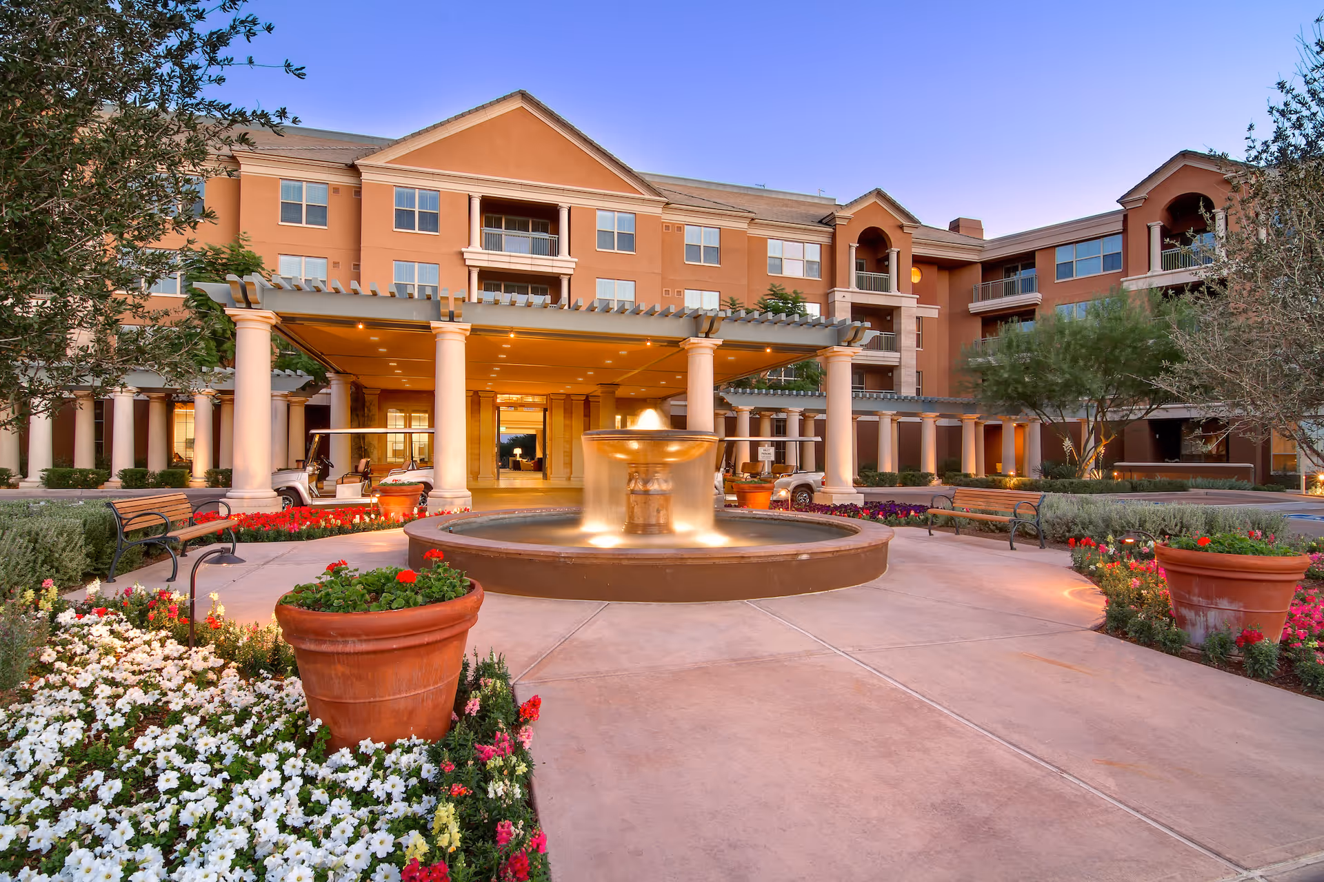 Front entrance of a multi-story senior living facility featuring a central illuminated fountain, columns, potted plants, benches, and flowerbeds.