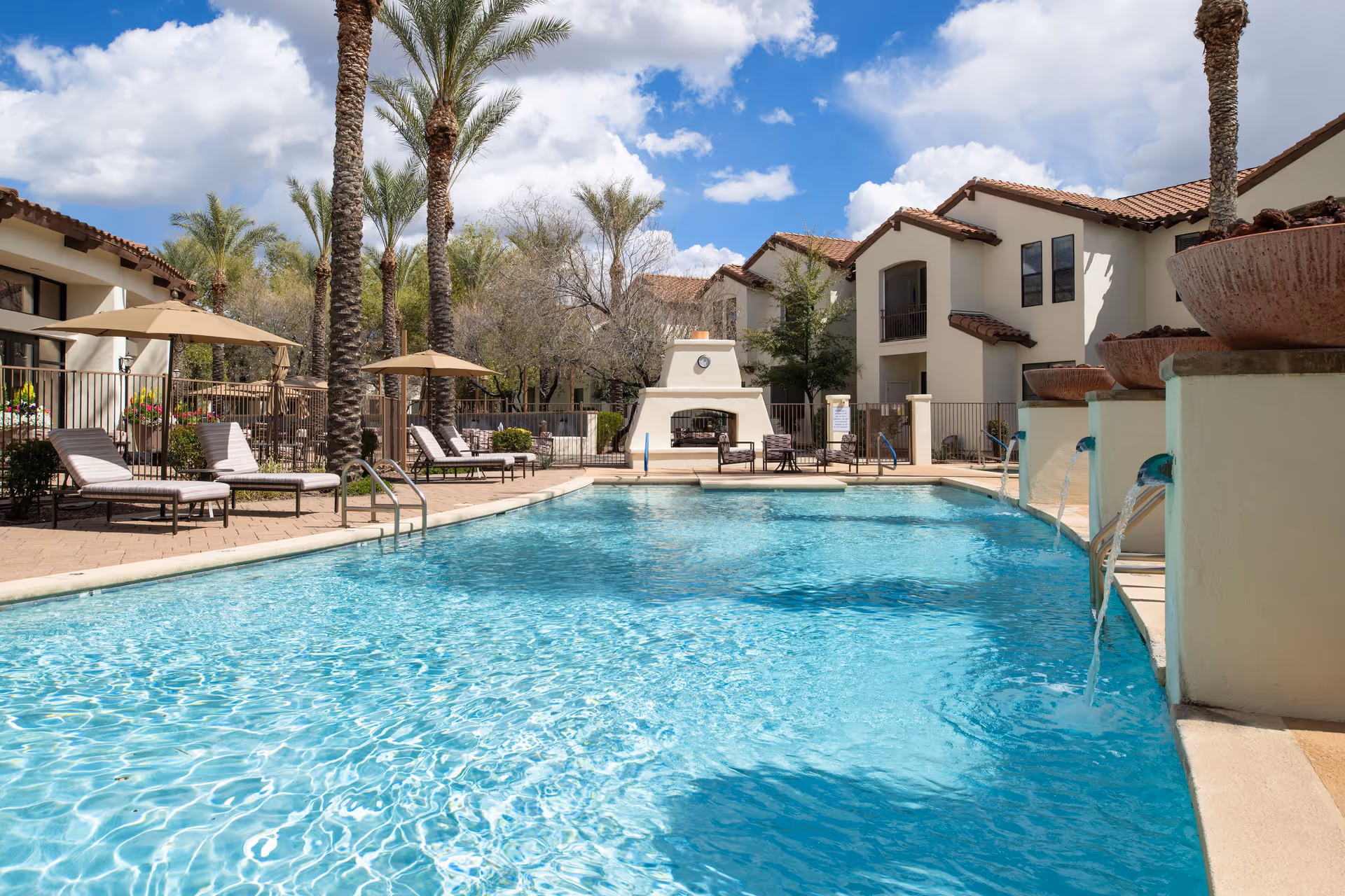 Outdoor swimming pool area at Villa Hermosa with clear blue water, several lounge chairs with umbrellas, palm trees, and a fireplace in the background under a partly cloudy sky.