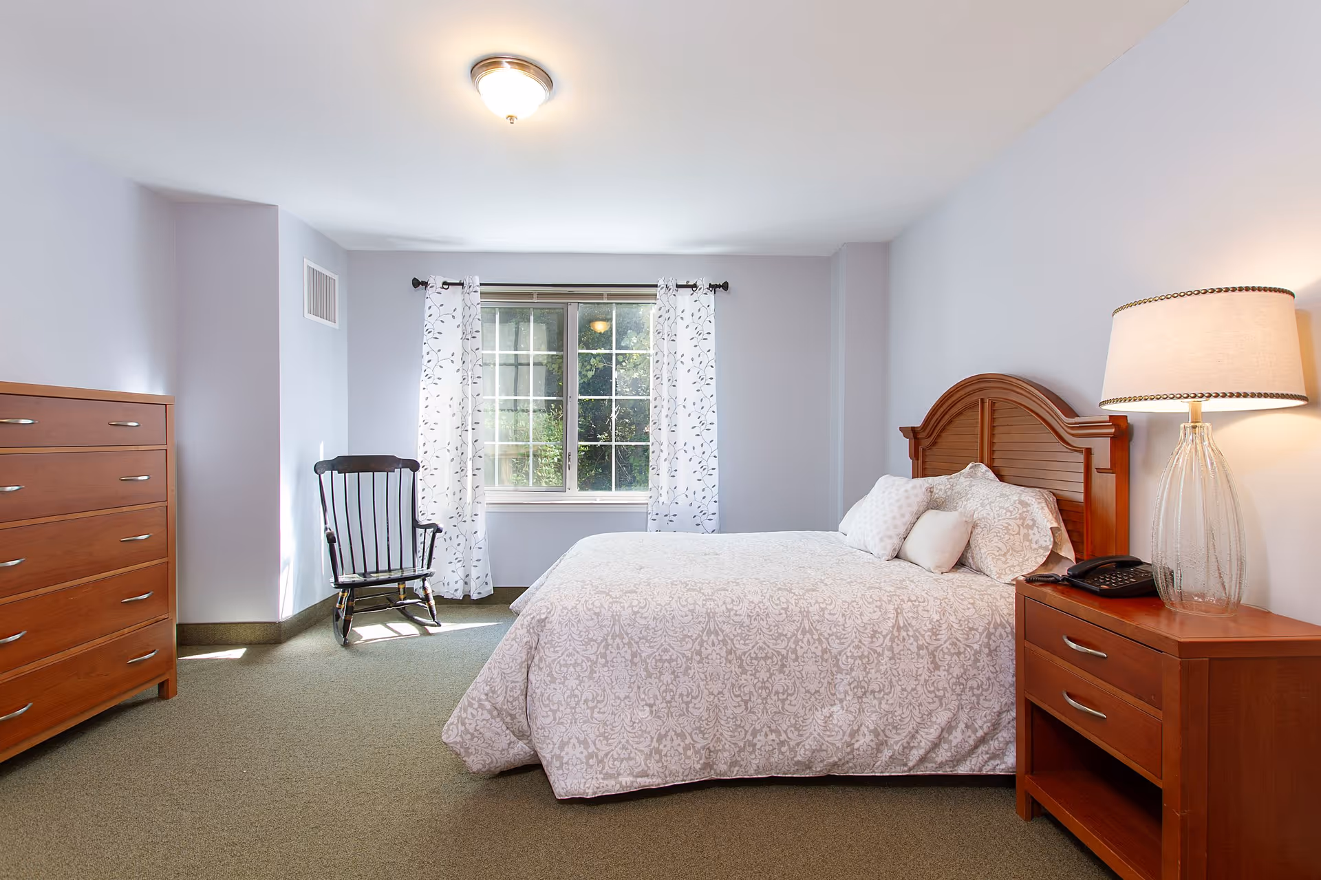 A bright bedroom with a large bed covered in a white patterned comforter and multiple pillows. To the right of the bed is a wooden nightstand with a glass lamp and a telephone. On the left side of the room is a wooden dresser and a black rocking chair near a window with white curtains featuring a subtle leaf pattern. The walls are painted light gray and the floor is carpeted in a muted green tone.
