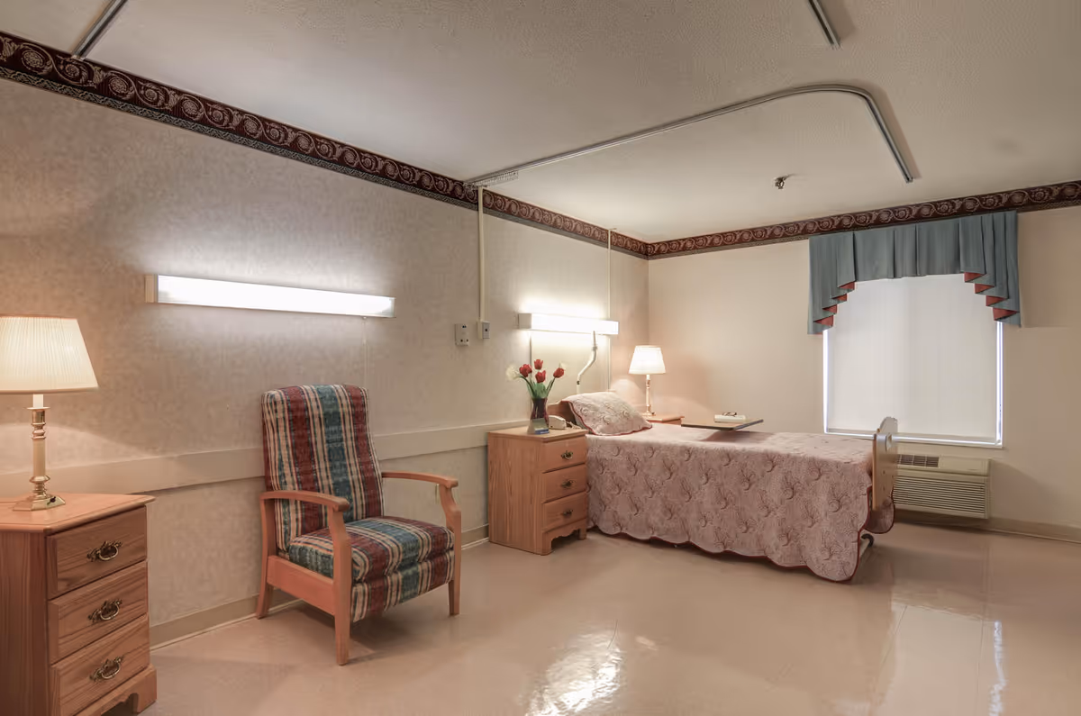 A simple, clean bedroom in a healthcare facility featuring a single bed with a pink quilted bedspread, two wooden nightstands with lamps, a wooden armchair with a striped cushion, a window with a blue valance, and a vase of red tulips on one nightstand.