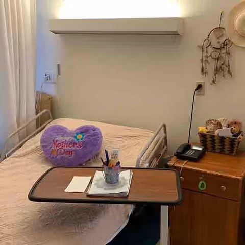 A senior living facility bedroom with a hospital-style bed covered in a beige blanket. On the bed is a purple heart-shaped pillow with the words 'Mother's Day' embroidered on it. In front of the bed is an overbed table holding a cup with pens and some papers. To the right of the bed is a wooden nightstand with a telephone, a basket containing dolls, and a dreamcatcher hanging on the wall above it.