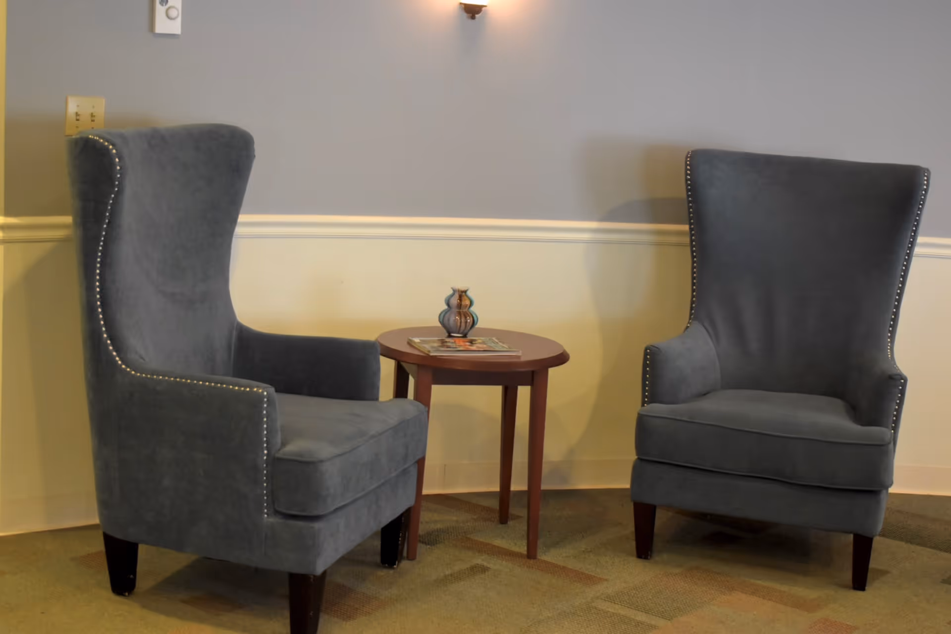 Two gray upholstered armchairs with nailhead trim facing each other with a small round wooden table between them. On the table is a small decorative vase and a magazine. The background shows a two-tone wall with a light fixture and electrical outlets.
