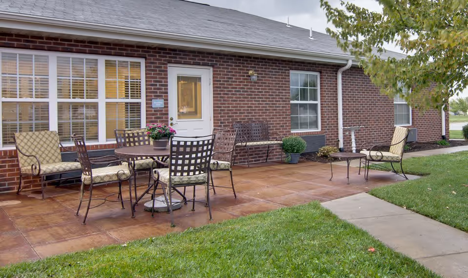 Outdoor patio area at Maple Tree Terrace with a round table and four cushioned chairs, two additional cushioned armchairs, a bench, potted plants, and a brick building with windows and a white door in the background.