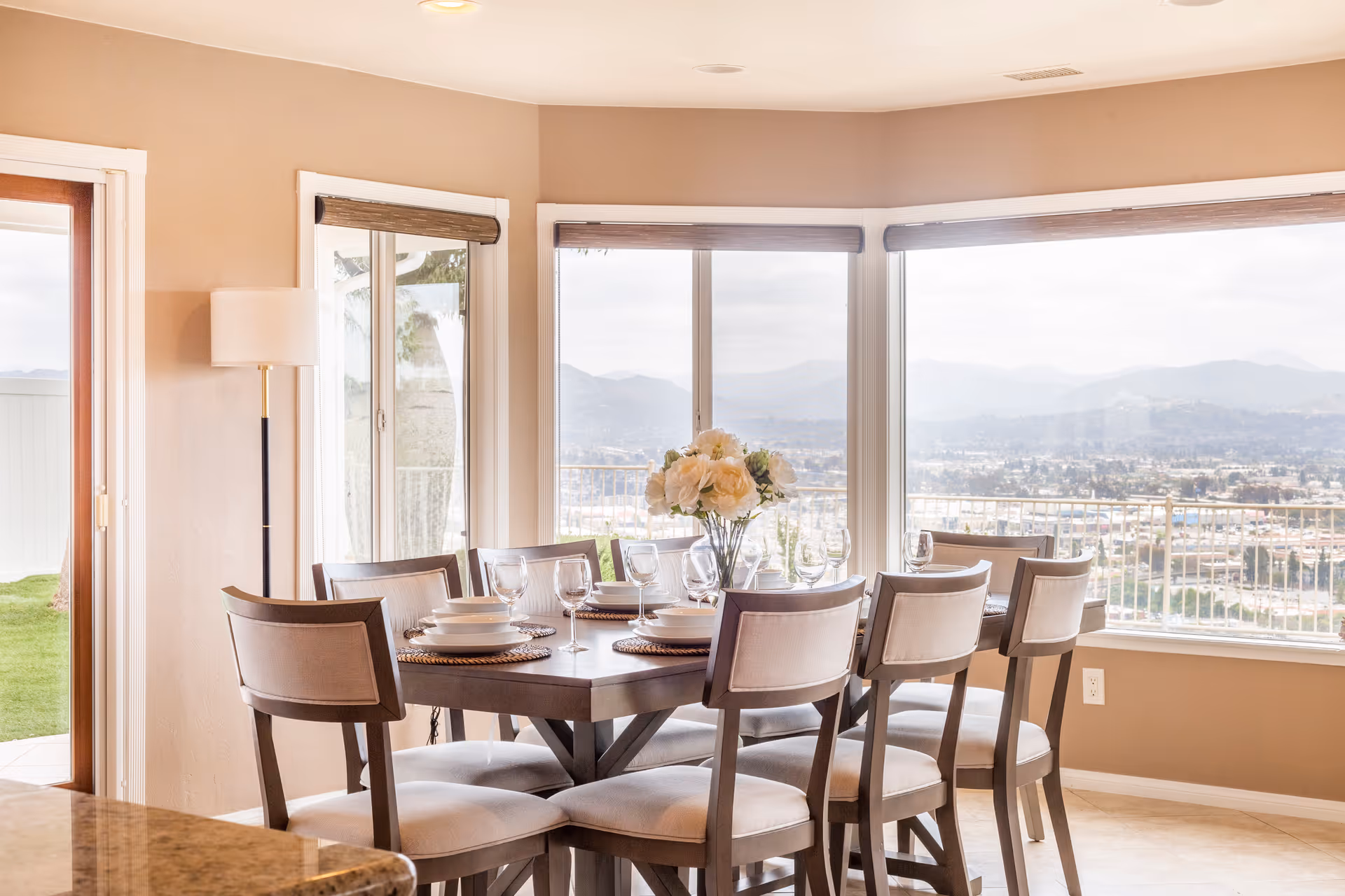 A bright dining room with a rectangular wooden table set for six, featuring white plates, bowls, and wine glasses. A vase with white flowers is centered on the table. Large windows provide a panoramic view of a cityscape and mountains in the distance. A sliding glass door opens to a grassy outdoor area.