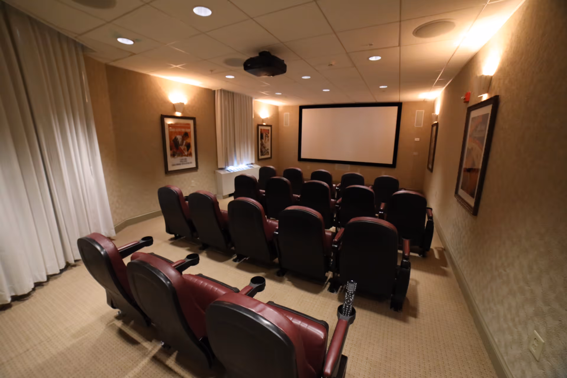 A small theater room with three rows of comfortable leather chairs facing a large blank projector screen. The room has beige walls with framed artwork, recessed ceiling lights, and white curtains covering the windows.