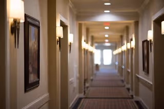 A long, well-lit hallway in an assisted living facility with wall-mounted lights and framed artwork on the walls. The carpeted floor has a striped pattern, and doors line both sides of the corridor leading to individual rooms.