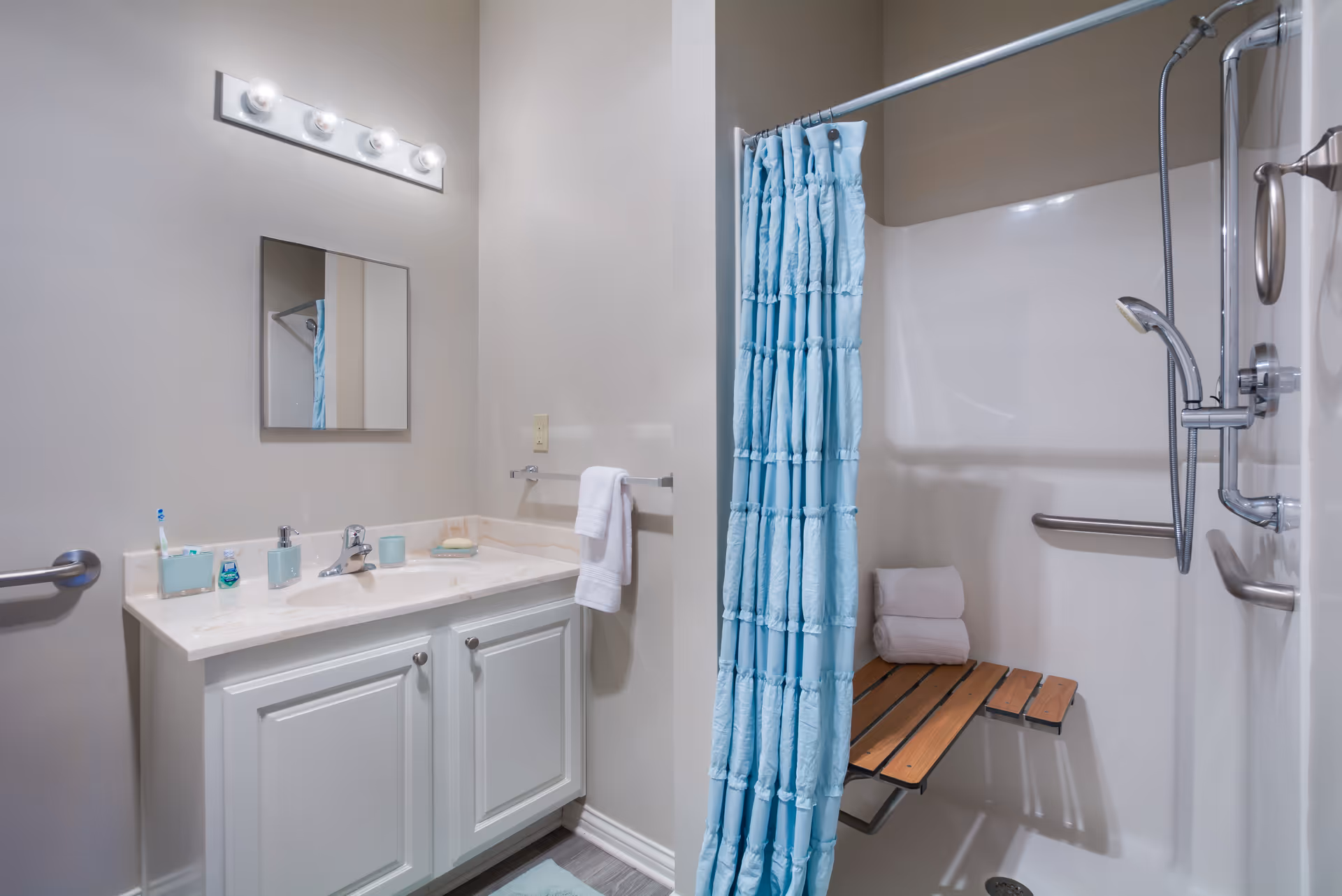 Bathroom with a vanity and mirror on the left and a shower with a blue curtain, fold-down wooden seat, and grab bars on the right.