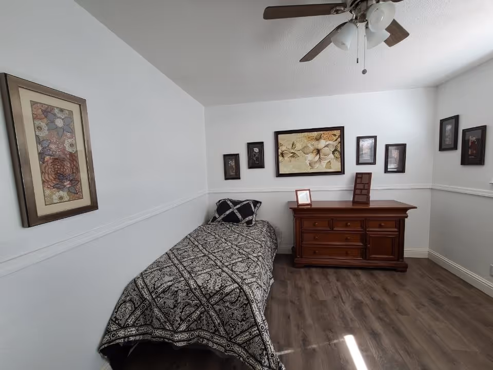A small bedroom with a single bed covered in a black and white patterned bedspread. There is a wooden dresser against the wall with framed pictures on top and several framed floral artworks hanging on the white walls. The room has wood flooring and a ceiling fan with lights.