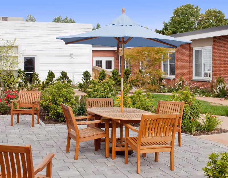 Wooden patio table and chairs with a blue umbrella on a paved courtyard surrounded by shrubs and brick and white buildings.