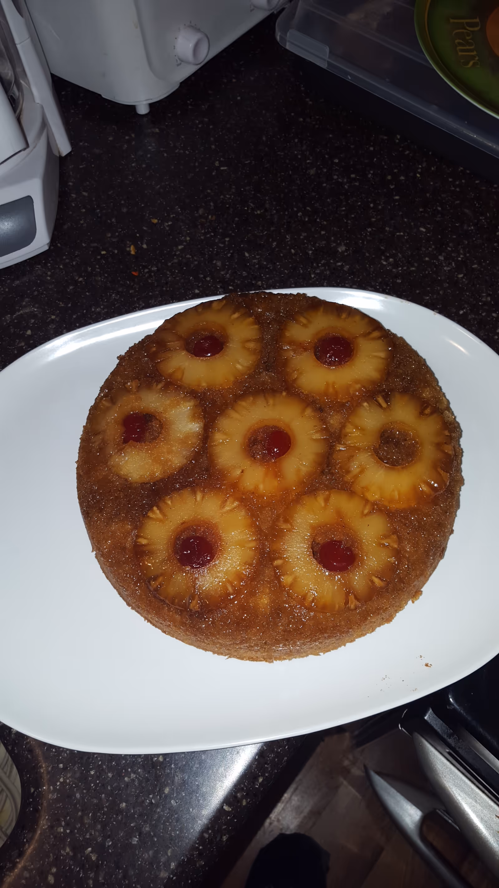 A pineapple upside-down cake on a white plate, topped with pineapple rings and maraschino cherries, placed on a dark kitchen countertop.
