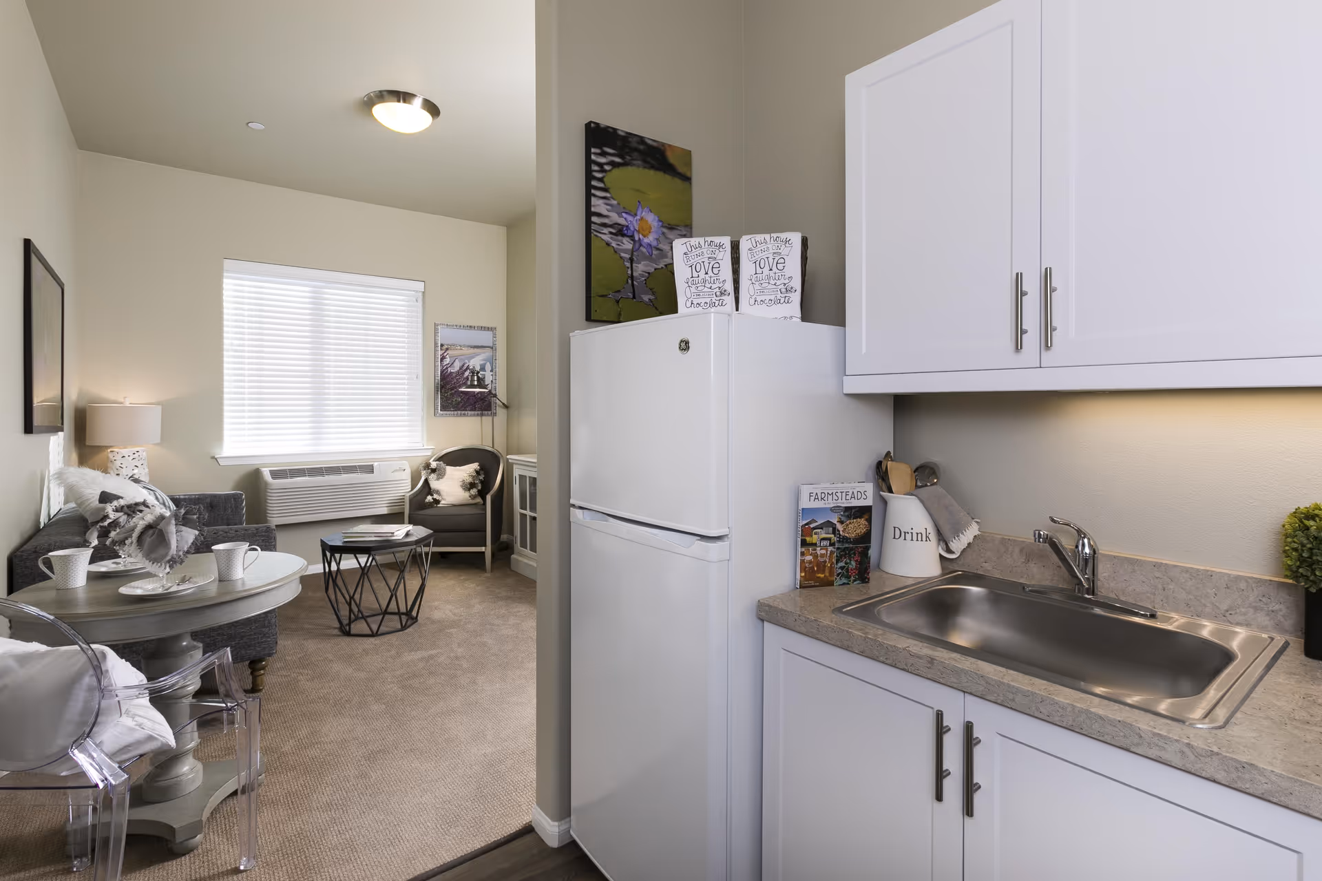 Interior view of a senior living facility apartment showing a small kitchen area with a white refrigerator, countertop with a sink, and white cabinets. Adjacent to the kitchen is a living room area with a gray sofa, a round table with two cups, a transparent chair, a small coffee table, an armchair, and a window with blinds letting in natural light.