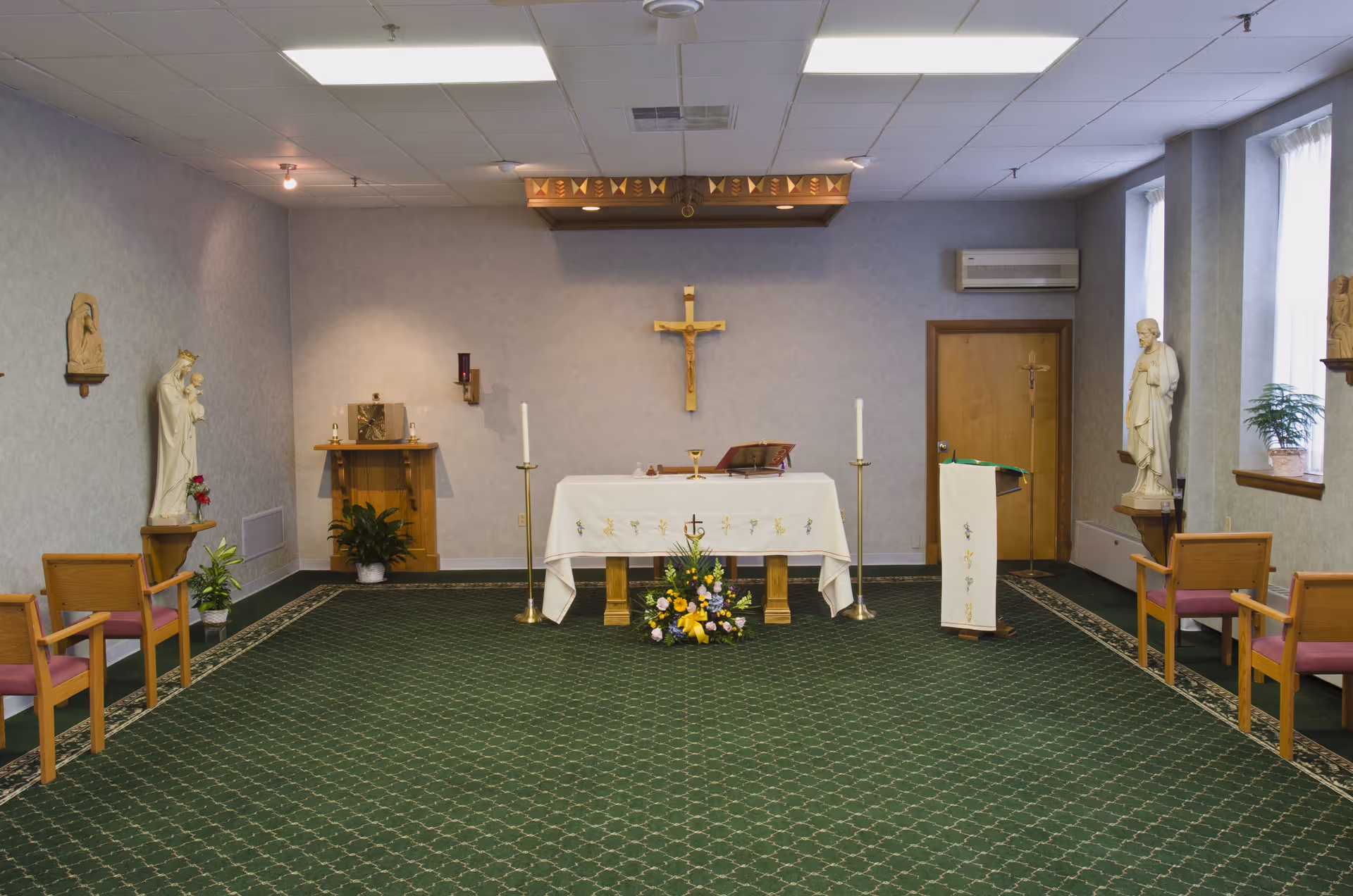 Interior of a small chapel with a green carpet, wooden chairs with pink cushions arranged along the sides, a white altar cloth on a wooden altar with candles and a flower arrangement in front. Religious statues and a crucifix are mounted on the walls.