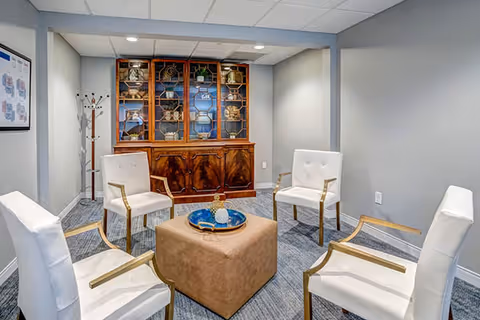 A small lounge with four white armchairs arranged around a central ottoman and a wooden display cabinet against a gray wall.