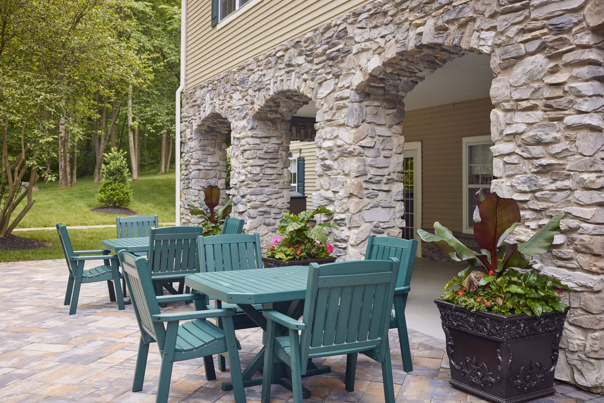 Outdoor patio area with green tables and chairs on a stone-paved surface next to a building with stone archways. There are large planters with green and red foliage plants, and a grassy area with trees in the background.