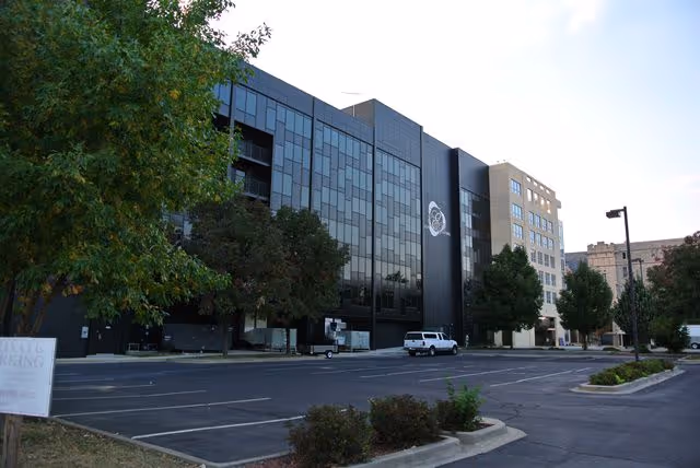 A modern multi-story building with a reflective glass facade, adjacent parking lot, and several trees.