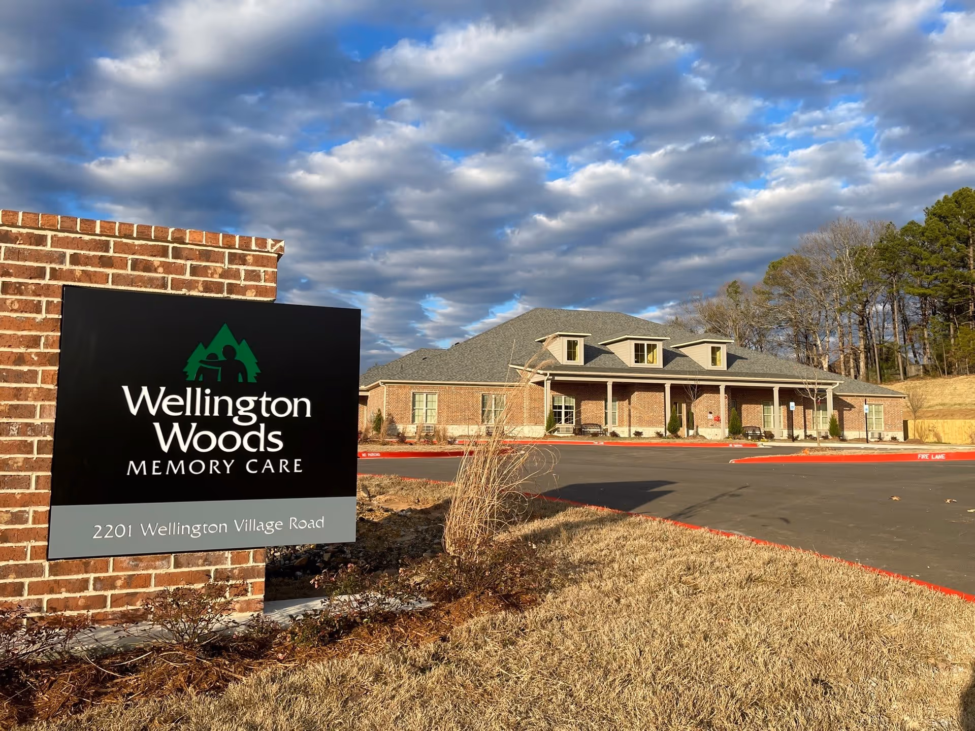 Exterior view of Wellington Woods Memory Care building with a brick sign in the foreground displaying the facility name and address, 2201 Wellington Village Road. The building is a single-story structure with a gray roof and several windows, surrounded by a parking lot and some landscaping under a partly cloudy sky.