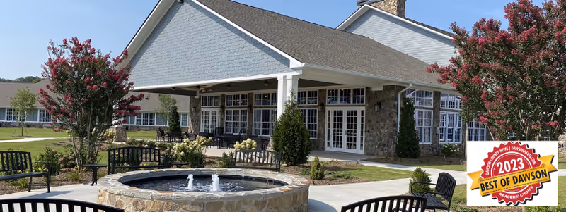 Outdoor view of Manor Lake Assisted Living, Memory Care, and Independent Living facility in Dawsonville, showing a stone water fountain surrounded by benches and landscaped greenery with flowering trees, and the building with large windows and a covered patio in the background under a clear blue sky.