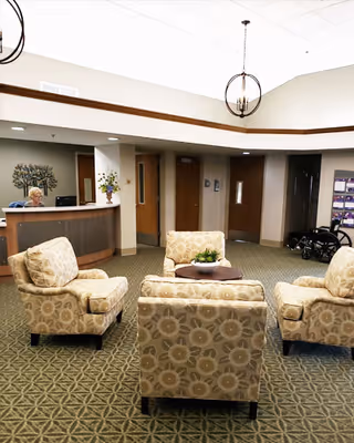 A seating area in a senior living facility with four patterned armchairs arranged around a small round table with a plant centerpiece. In the background, there is a reception desk with a staff member, doors, a wheelchair, and a bulletin board on the wall. The ceiling has a modern light fixture and the floor is carpeted with a geometric pattern.