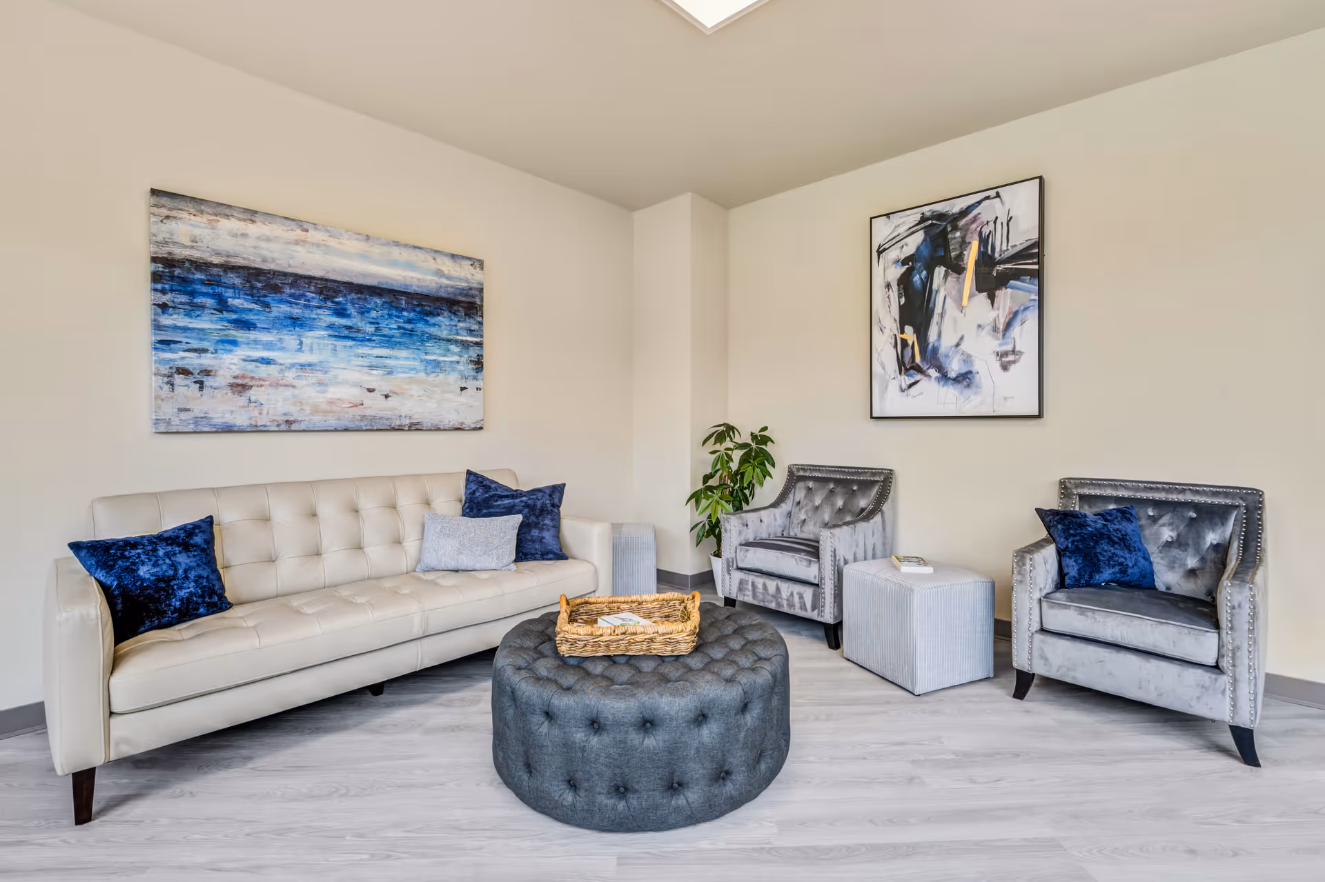 Bright seating area with a tufted cream sofa, two gray armchairs, a round tufted ottoman and artwork on the walls.