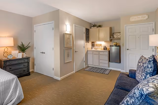 A cozy interior room featuring a small kitchenette with cabinets, a mini refrigerator, and a countertop with a lighted 'cheers' sign. To the left, there is a black dresser with a lamp, clock, and plant on top. The room has beige walls, two closed white doors, framed artwork, and a blue couch with patterned pillows on the right side.