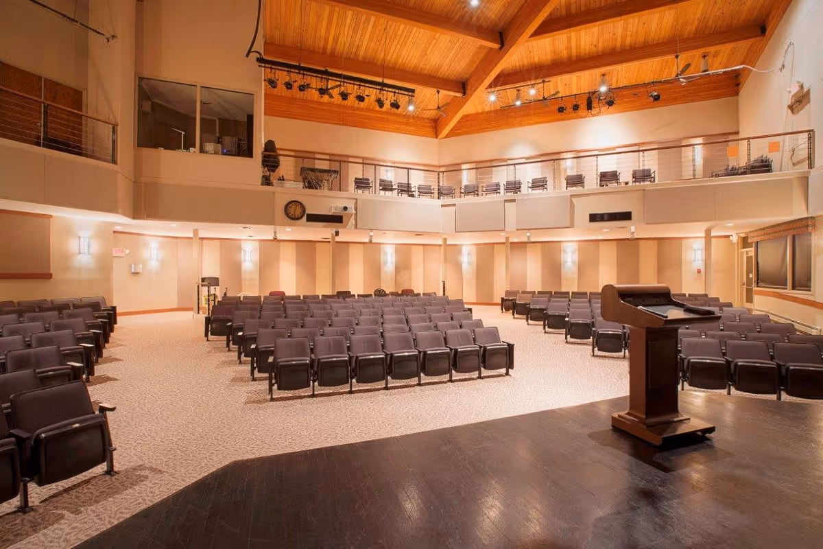 Interior view of a large auditorium or lecture hall with rows of empty chairs facing a wooden podium on a stage. The room has a high wooden ceiling with exposed beams and lighting fixtures. There is a balcony area with additional seating and a clock on the wall above the main seating area.