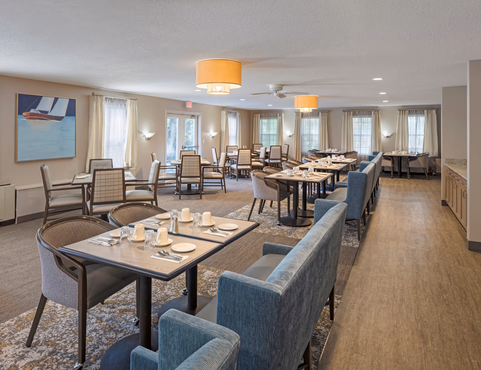 A spacious dining room with multiple tables set for meals, featuring chairs and cushioned bench seating. The room has large windows with curtains, soft lighting from ceiling fixtures, and a painting of a sailboat on one wall. The floor is a combination of carpet and wood, and there is a counter with cabinets along one side.