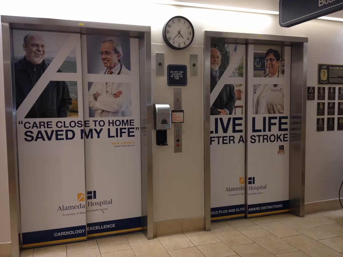 Two elevator doors inside Alameda Hospital with large posters. The left elevator shows a smiling man and a doctor with the text 'CARE CLOSE TO HOME SAVED MY LIFE' and 'Alameda Hospital Cardiology Excellence'. The right elevator shows a man and a female doctor with the text 'LIVE LIFE AFTER A STROKE' and 'Alameda Hospital Gold Plus and Elite Award Distinctions'. A clock and elevator buttons are visible between the elevators.