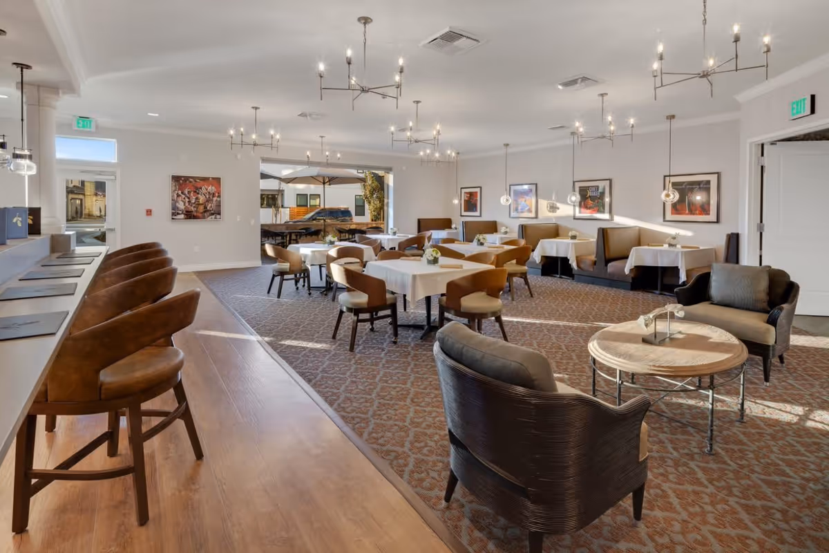 A spacious dining room with several tables covered with white tablecloths, surrounded by chairs and booth seating along the wall. There are modern chandeliers and pendant lights hanging from the ceiling. On the left side, there is a counter with high-back chairs. The room has large windows and doors letting in natural light, and framed artwork on the walls.
