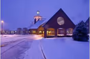 Exterior view of a building at dusk with snow covering the ground and a lit pathway leading to the entrance. The building has a peaked roof with a circular window and a small tower with a light on top. A snow-covered tree is visible on the right side.