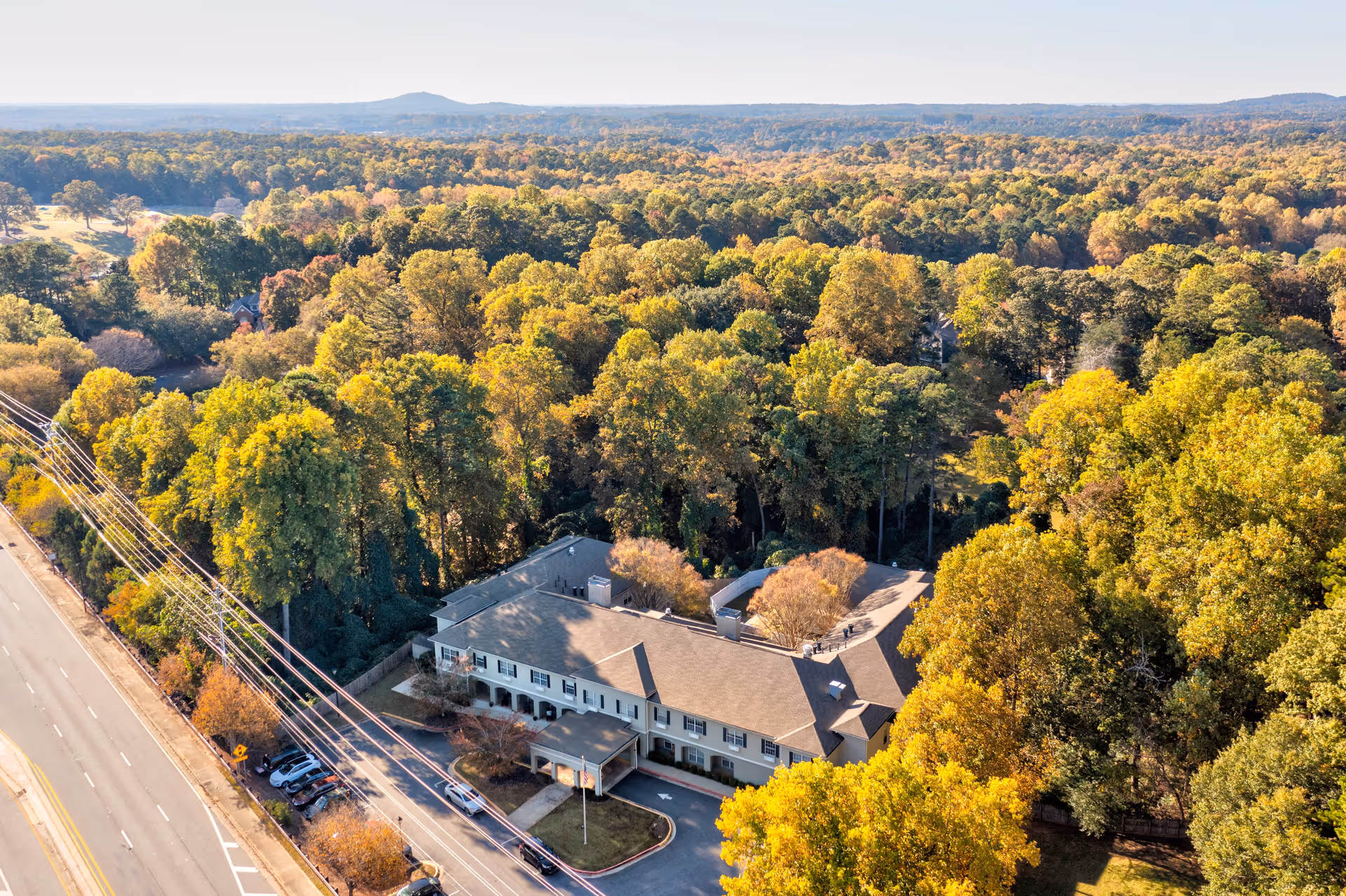 Aerial view of Greenwood Place Assisted Living and Memory Care facility surrounded by dense trees with autumn foliage, located next to a road with several parked cars and power lines visible.
