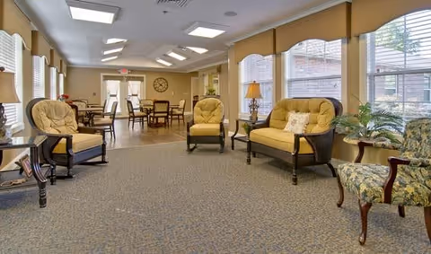A bright and spacious senior living common area with large windows letting in natural light. The room features cushioned armchairs and a loveseat arranged around small side tables, with additional dining tables and chairs visible in the background. The decor includes a table lamp, potted plants, and a wall clock, creating a warm and inviting atmosphere.