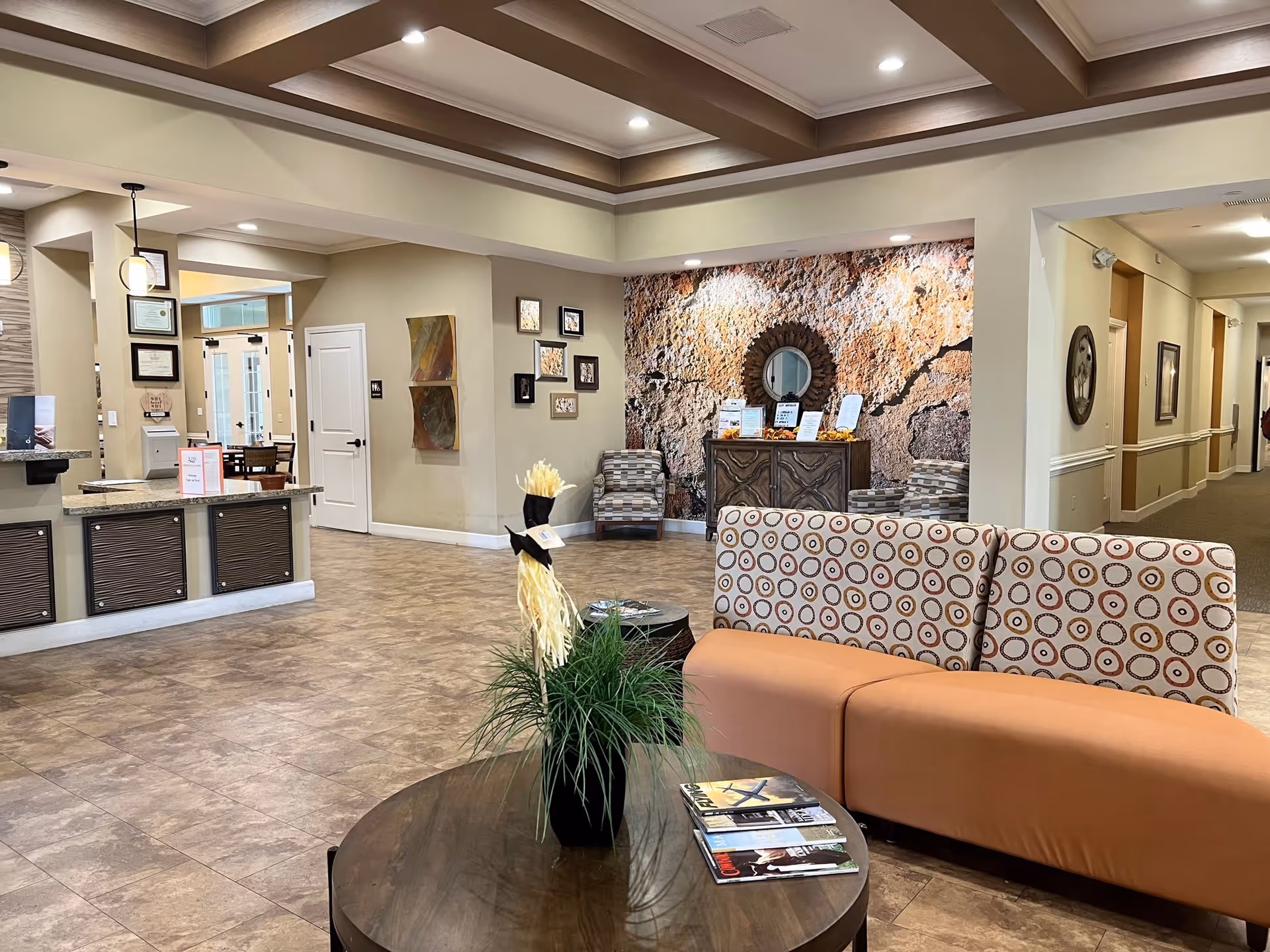 A spacious and well-lit senior living facility common area with tiled floors and a coffered ceiling. The room features a patterned sofa, a round wooden coffee table with a plant and magazines, and two armchairs near a textured wall with a circular mirror above a wooden cabinet. The area also includes a reception desk on the left and a hallway leading to other rooms on the right.