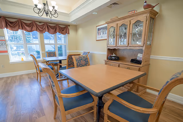 Small dining area with wooden tables and upholstered chairs, a china cabinet, and windows with valances.