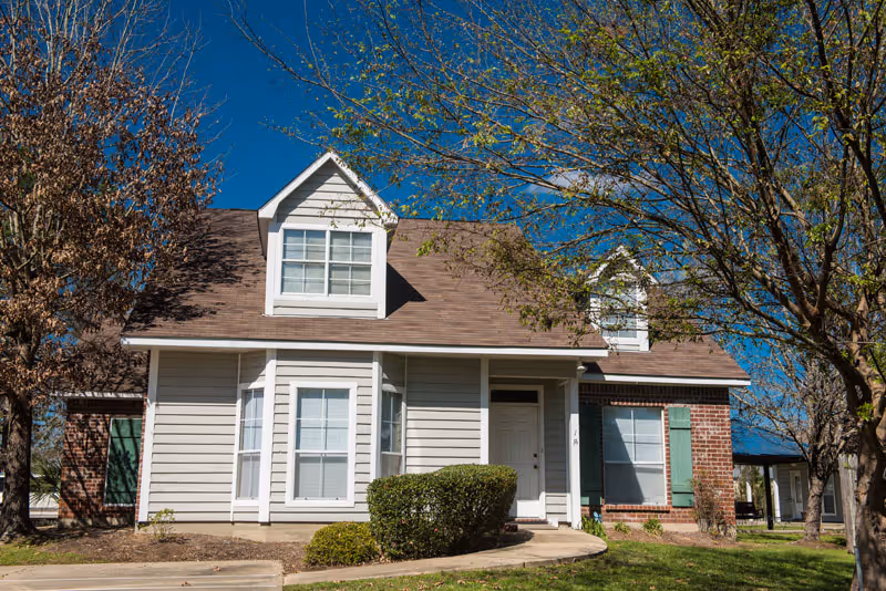 Exterior view of a single-story residential building with beige siding and brick accents, featuring a brown shingled roof with dormer windows. The front entrance has a white door and is surrounded by green bushes and trees under a clear blue sky.