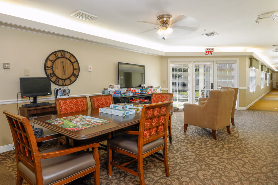 A cozy common area in an assisted living facility featuring a wooden table with six chairs, some with red patterned cushions. On the table are several board games and puzzles. Behind the table is a TV mounted on the wall with more board games stacked below it. A large wooden clock hangs on the wall to the left. There is a computer on a small desk in the corner. Two beige armchairs face the TV. The room has beige walls, patterned carpet, and a ceiling fan with lights. French doors with blinds lead to an outdoor area, and a hallway extends to the right.