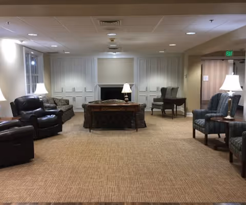 Spacious senior living facility lounge with armchairs, sofas, side tables and lamps facing built-in cabinets and a fireplace.