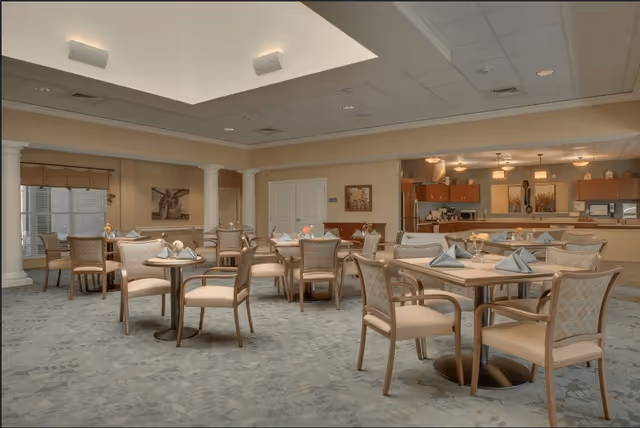 A spacious dining room with multiple tables and chairs arranged neatly. Each table is set with folded napkins and small floral centerpieces. The room features a high ceiling with a skylight, soft lighting, and a kitchen area visible in the background with wooden cabinets and stainless steel appliances.