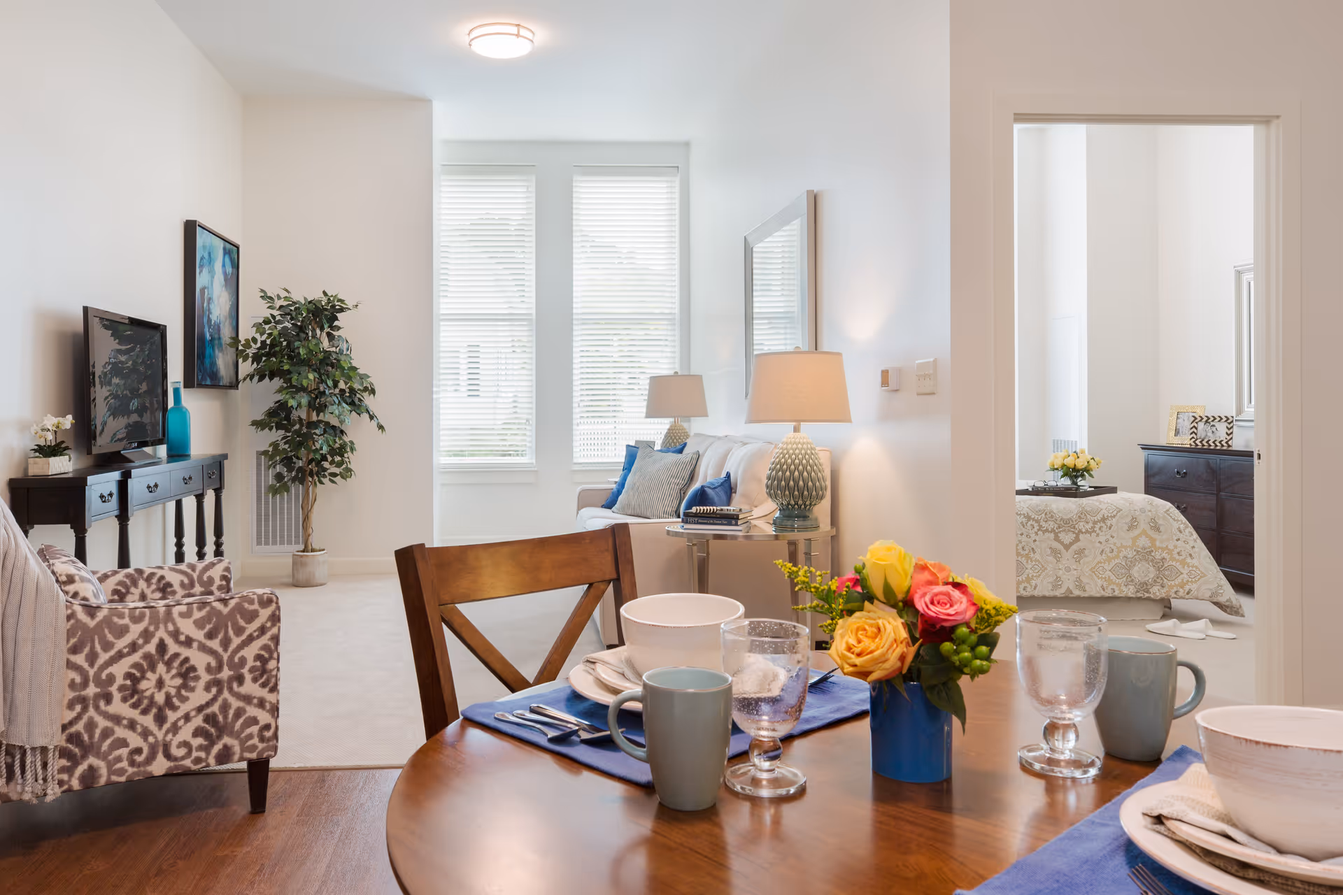 A bright and cozy living and dining area in a senior living facility. The dining table is set with plates, bowls, cups, and glasses, and decorated with a small bouquet of colorful flowers. The living room features a patterned armchair, a sofa with blue and gray cushions, a side table with a lamp, a TV on a console table, and a large potted plant near two tall windows with blinds. A doorway leads to a bedroom with a bed, dresser, and framed pictures.