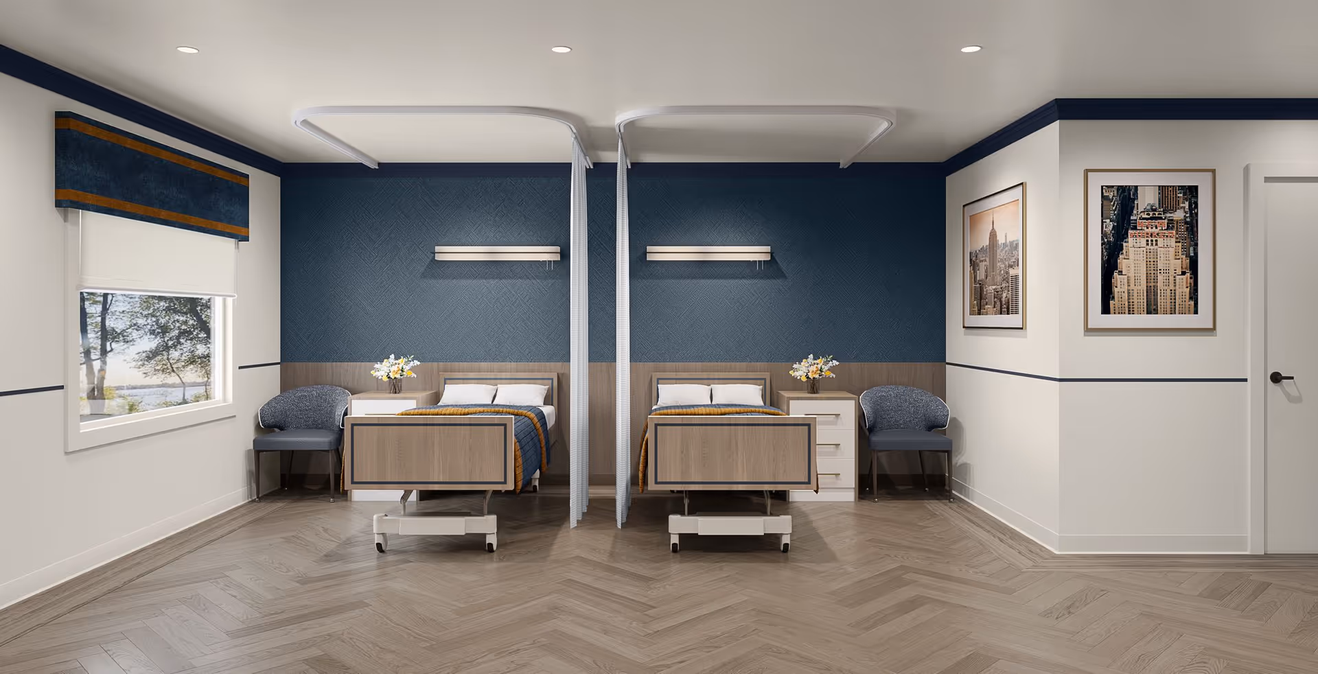 A clean and modern nursing facility room with two hospital beds separated by a privacy curtain. Each bed has a bedside table with a vase of flowers and a chair next to it. The walls are painted white with a dark blue accent wall behind the beds. There are two framed pictures on the right wall and a window with a blue and orange valance on the left. The floor has a light wood herringbone pattern.
