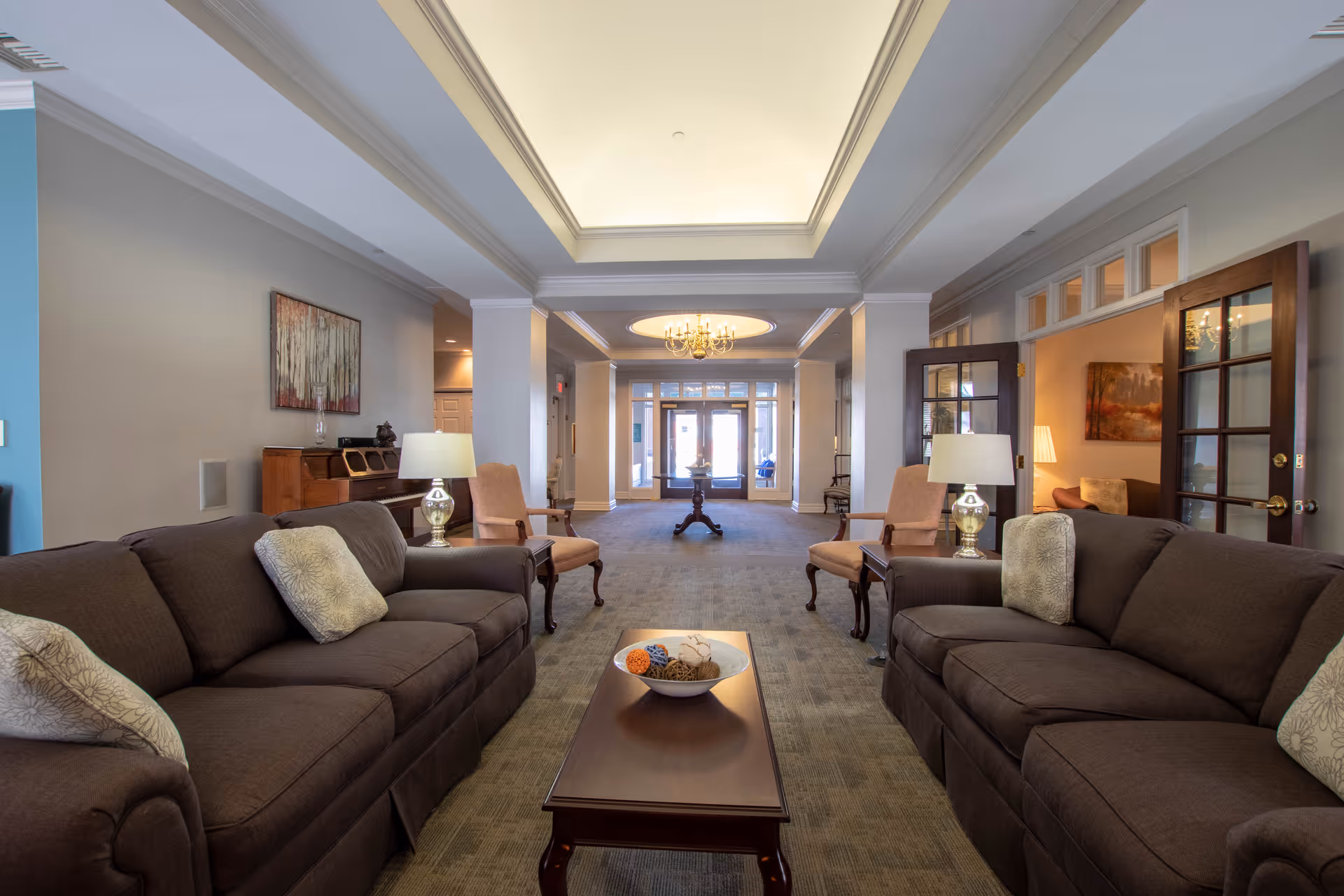 A spacious and well-lit living room area in a senior living facility with two brown sofas facing each other, a wooden coffee table with a decorative bowl in the center, two armchairs with side tables and lamps, a piano against the left wall, and a chandelier hanging from the ceiling near the entrance doors.