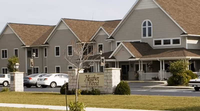 Exterior view of HeatherWood Assisted Living & Memory Care facility showing a large beige building with multiple peaked roofs, several windows, a covered porch area, a parking lot with cars, and a sign in front with the facility name.