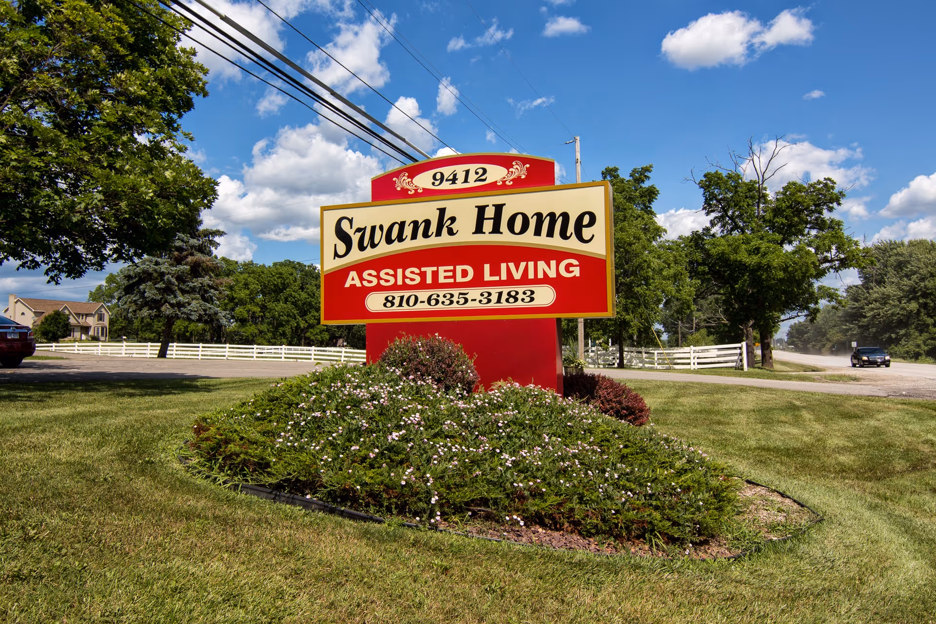 A large red and cream sign for Swank Home Assisted Living with the phone number 810-635-3183, situated on a grassy area with bushes and flowers. Trees, a road, and a house are visible in the background under a partly cloudy blue sky.