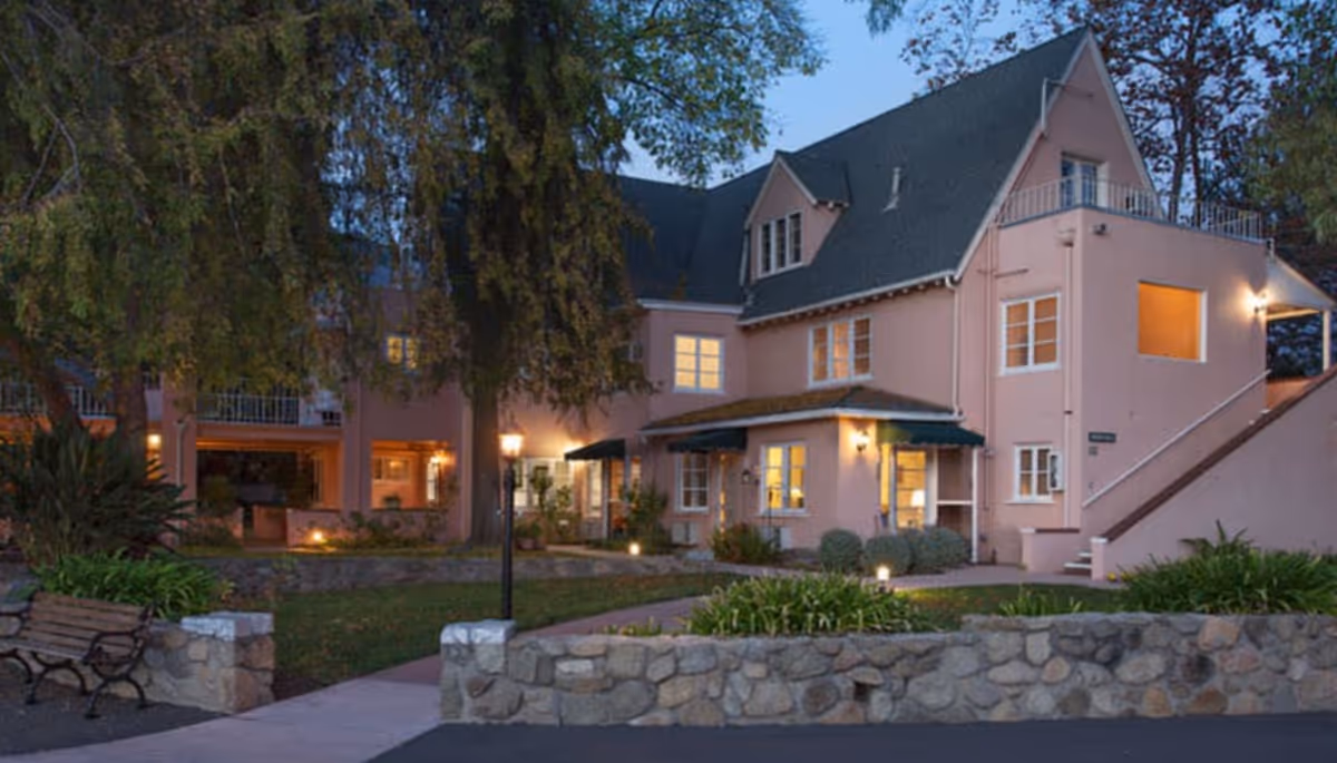 Two-story pink residential building with a landscaped courtyard, stone walls, benches, and exterior lighting at dusk.