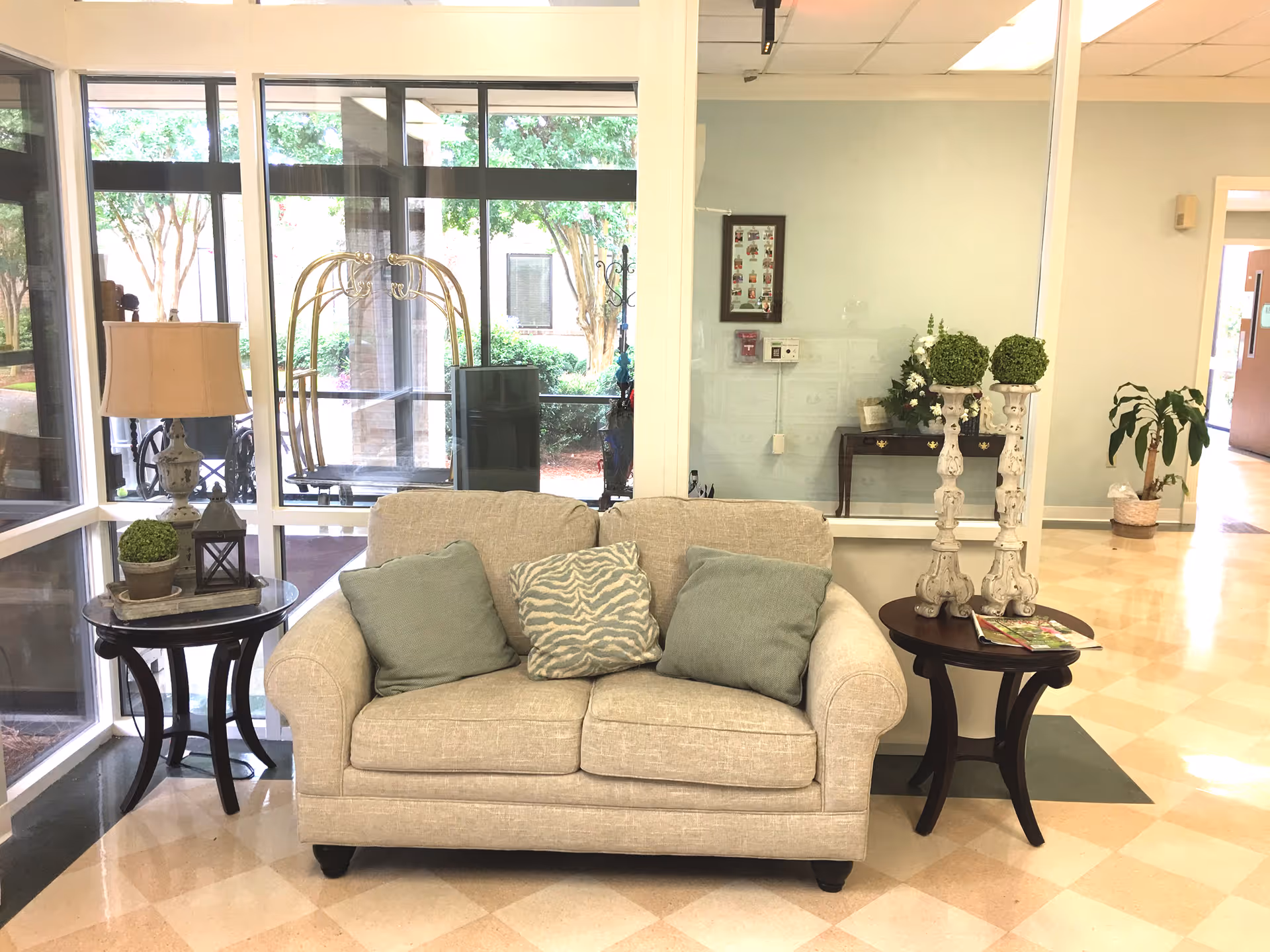 A cozy seating area in a healthcare facility lobby featuring a beige loveseat with three cushions, flanked by two dark wooden side tables. One table holds a lamp, a small potted plant, and a lantern, while the other has two decorative white candle holders with green topiary balls and some magazines. Large windows behind the loveseat reveal an outdoor view with trees and a luggage cart.