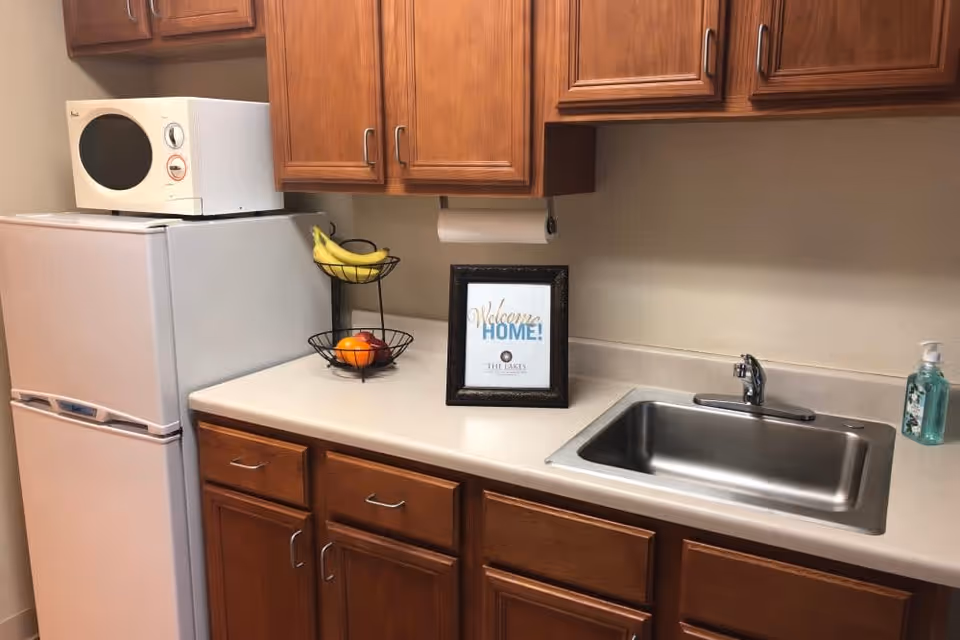 A small kitchen area with wooden cabinets, a white refrigerator with a microwave on top, a two-tier fruit basket containing bananas and oranges, a framed sign that says 'Welcome Home! The Lakes', a stainless steel sink, and a bottle of hand soap on the countertop.