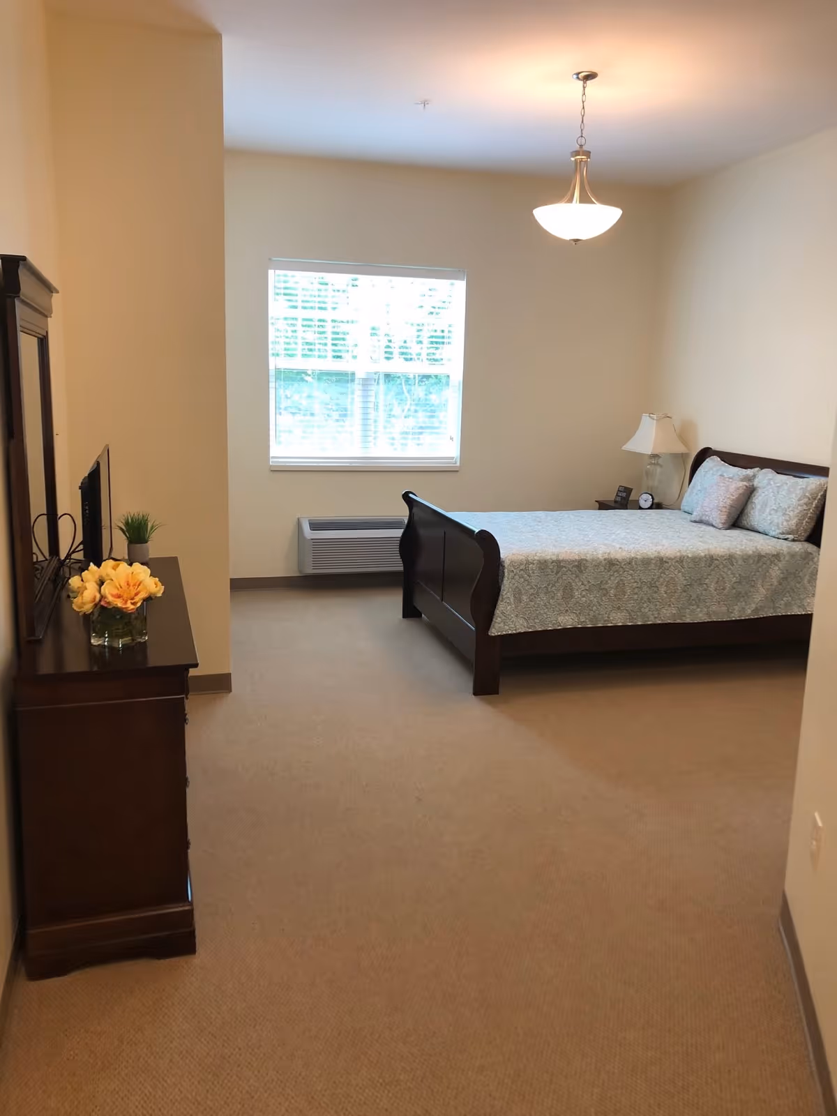 A bedroom with a wooden bed frame and a neatly made bed with patterned bedding. There is a nightstand with a lamp and an alarm clock next to the bed. A window with blinds is on the far wall, and a wooden dresser with a mirror and decorative flowers is on the left side of the room. The room has beige walls and carpeted floor.
