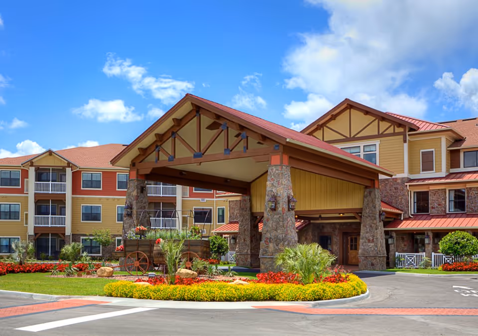 Front exterior view of a senior living facility named American House Wildwood with a large covered entrance supported by stone pillars, surrounded by well-maintained landscaping including colorful flowers and greenery under a blue sky with some clouds.