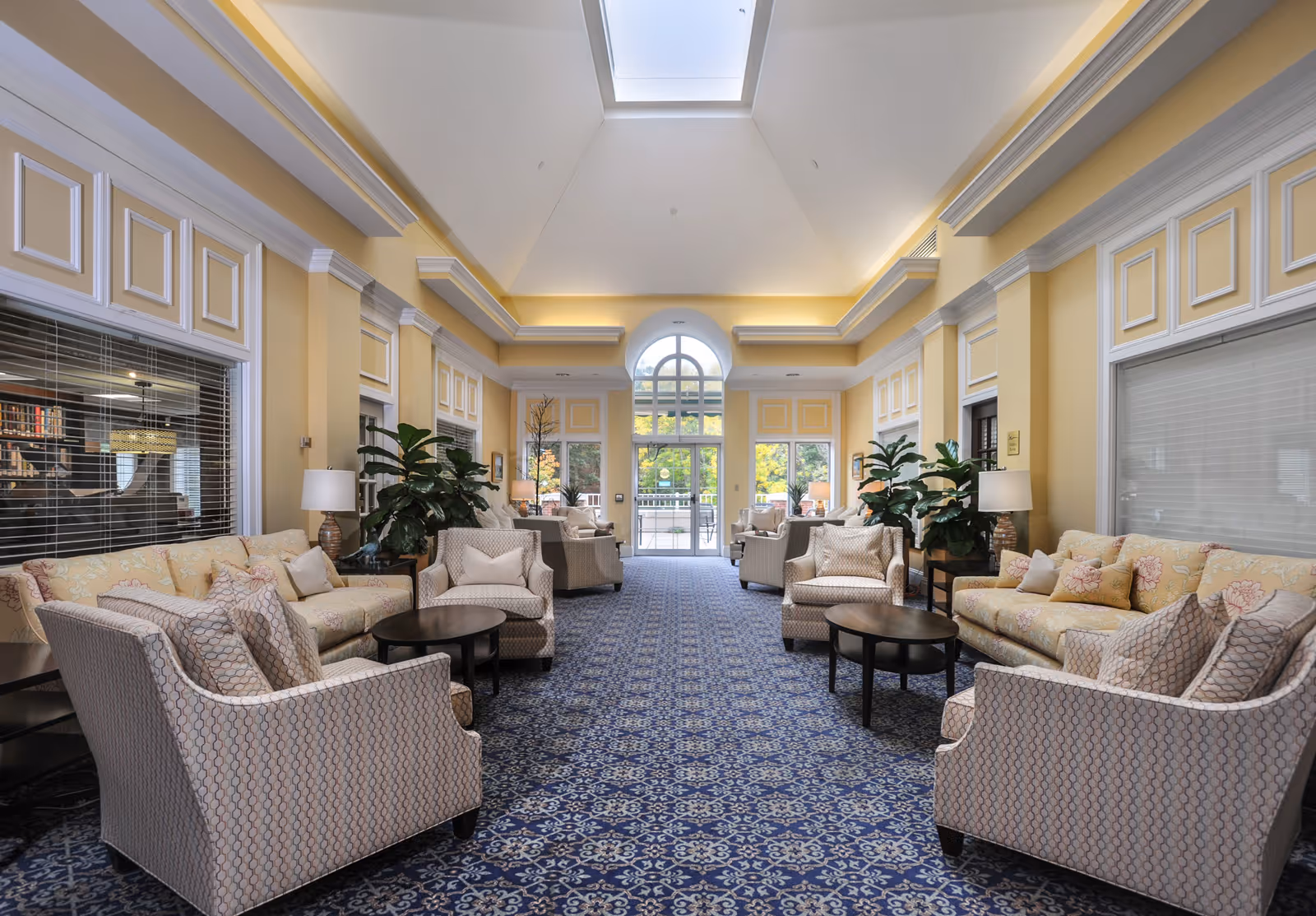 A spacious and elegant living room area with patterned blue carpet, beige and floral upholstered sofas and armchairs arranged around dark wooden coffee tables. The room has high ceilings with a skylight, large windows, yellow walls with white trim, and several green potted plants. There is a glass door at the far end leading outside.