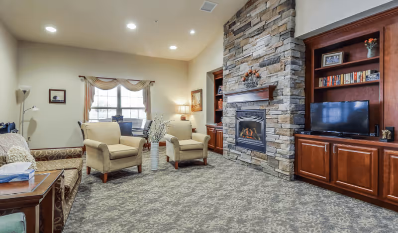 A cozy living room with a stone fireplace in the center, flanked by wooden built-in shelves and cabinets. Two beige armchairs and a patterned sofa are arranged around the fireplace. A window with curtains lets in natural light, and a floor lamp and table lamp provide additional lighting. The carpet has a subtle floral pattern.