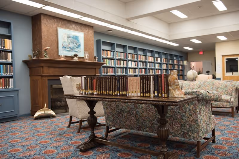 A cozy library room with a wooden table in the center holding a row of books. Behind the table is a floral-patterned sofa and a cushioned armchair. The room features a wooden fireplace with decorative items on the mantel, blue built-in bookshelves filled with books, and a patterned carpet. The ceiling has recessed lighting, and there is a globe on a side table near another sofa in the background.
